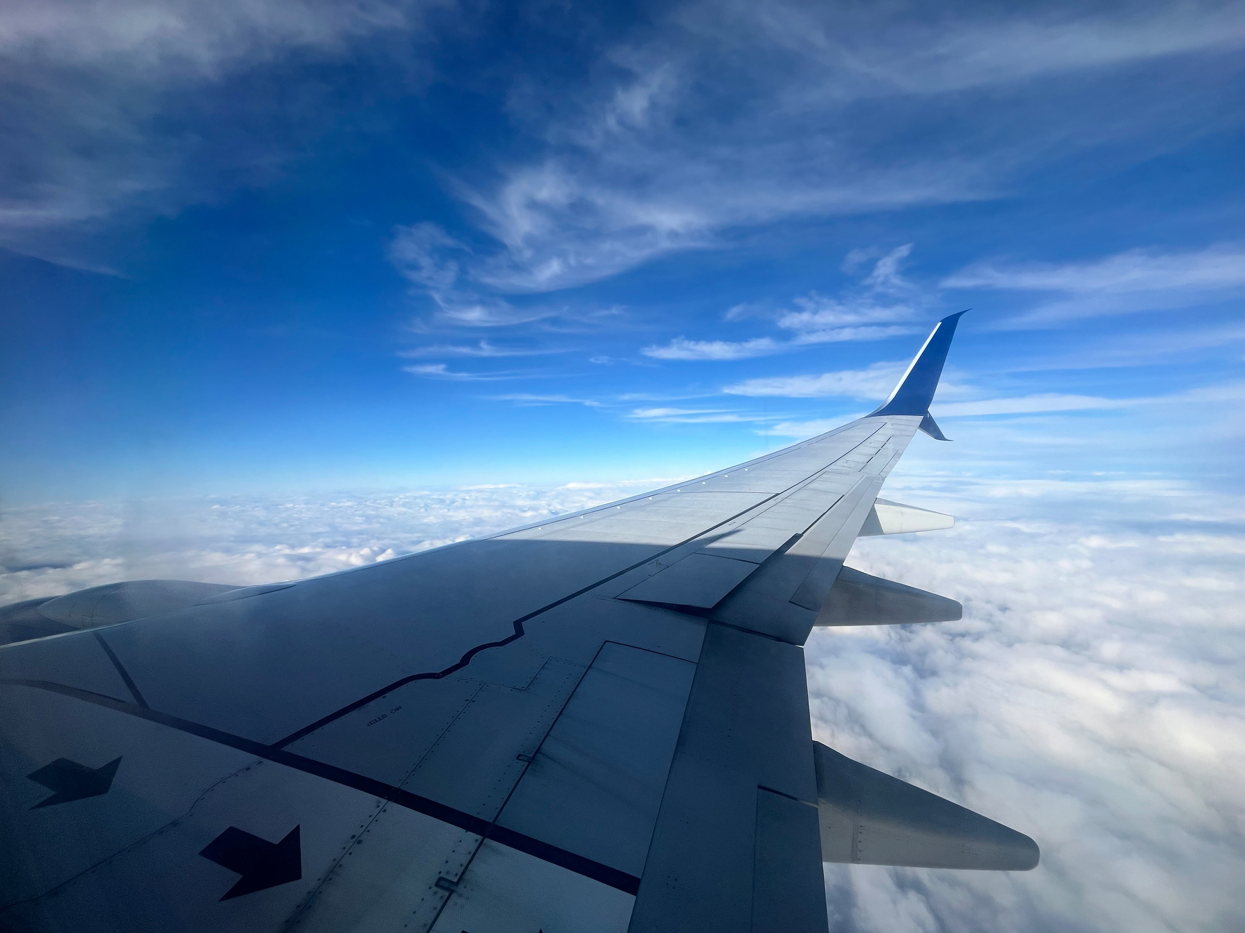 An airplane wing is seen from a window, soaring above a blanket of clouds under a clear blue sky.