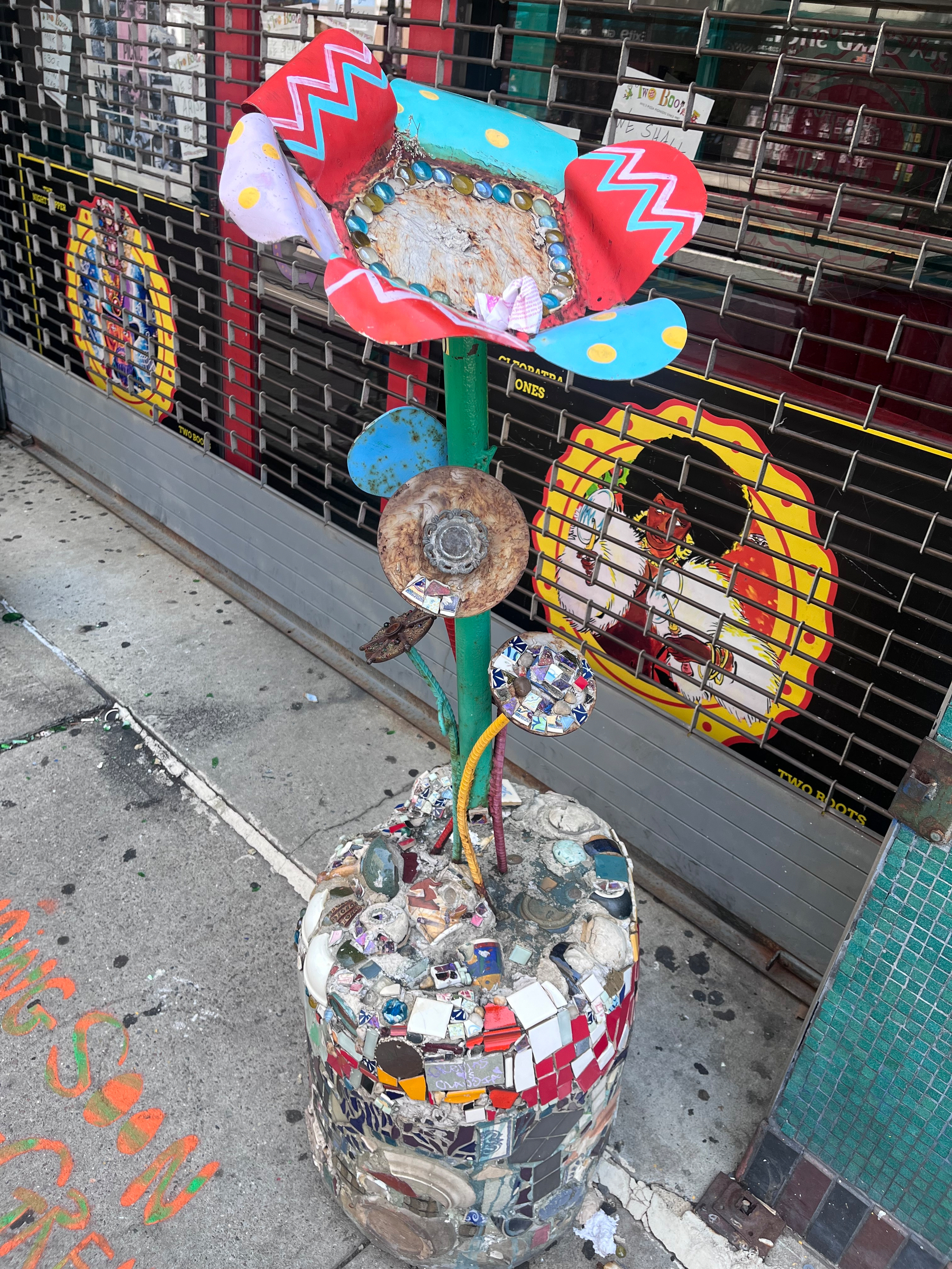 A colorful, mosaic-style flower sculpture made from various materials stands on a sidewalk in front of a closed storefront with decorative artwork on the metal gate.