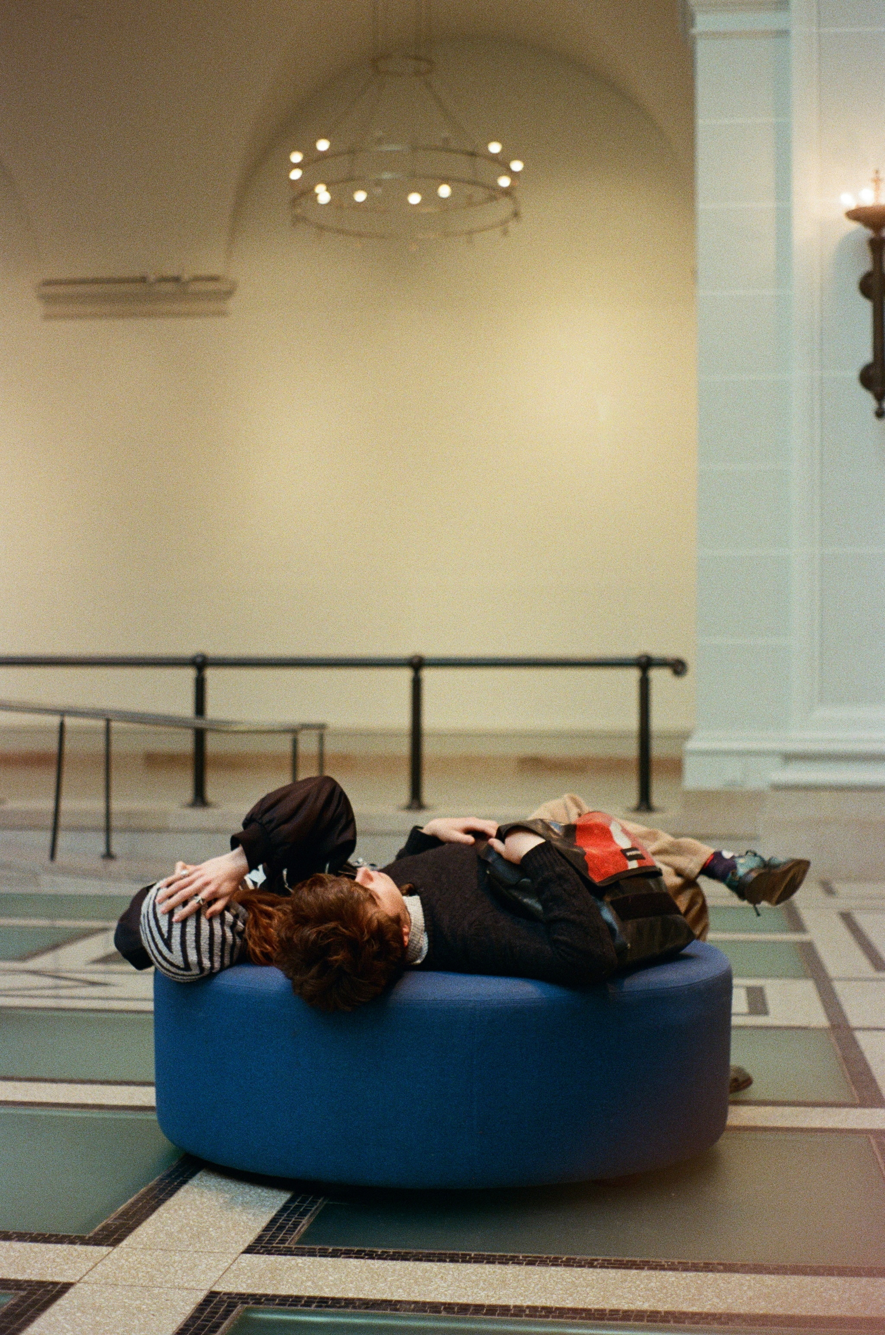 Two people are lying on a blue circular bench inside a spacious room with a chandelier.