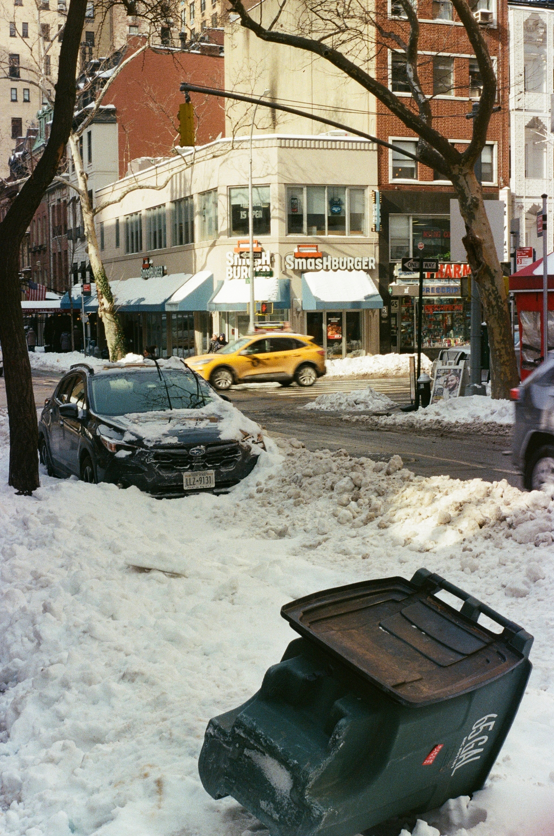 A snow-covered street with a parked car and overturned trash bin, surrounded by buildings including a Smashburger restaurant and a passing yellow taxi.